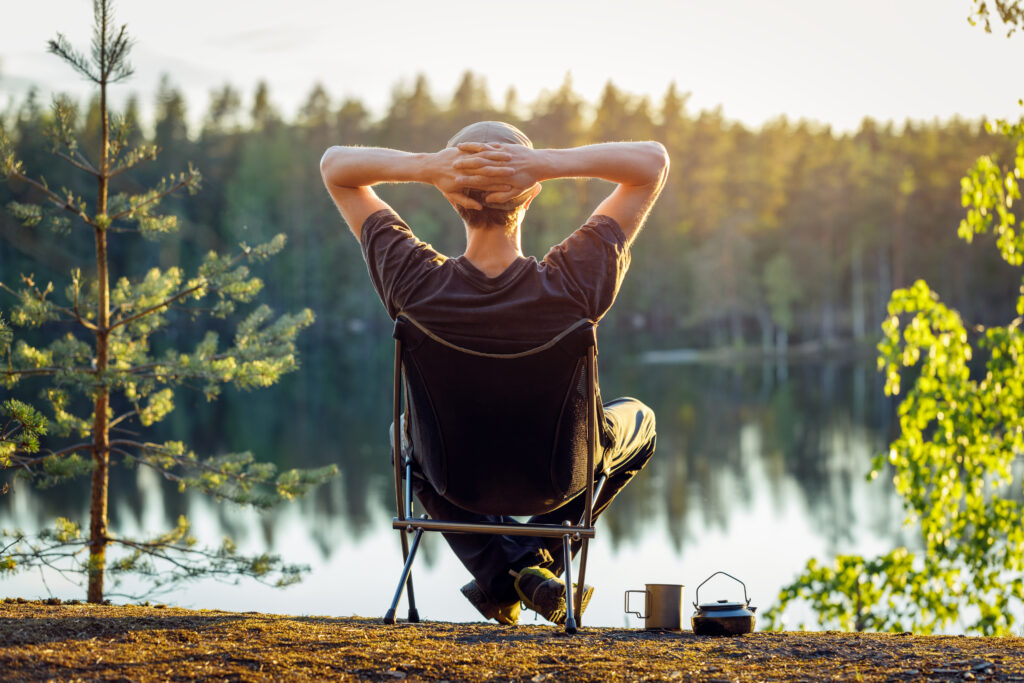 Man Is Sitting In A Camping Chair On The Background Of A Forest Lake On A Beautiful Summer Evening.
